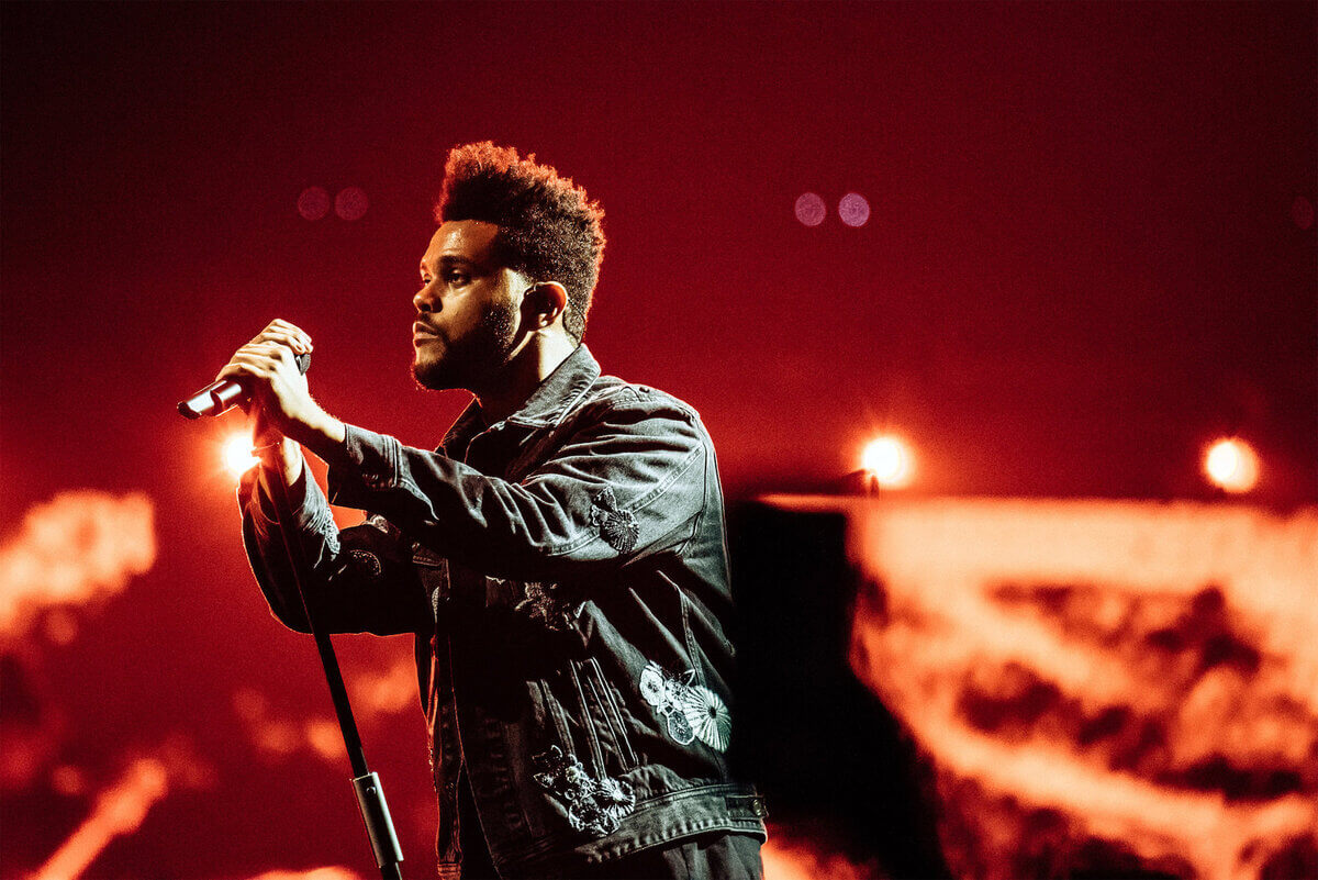 The Weeknd mengenakan jaket jeans sambil memegang mic sedang tampil di atas panggung